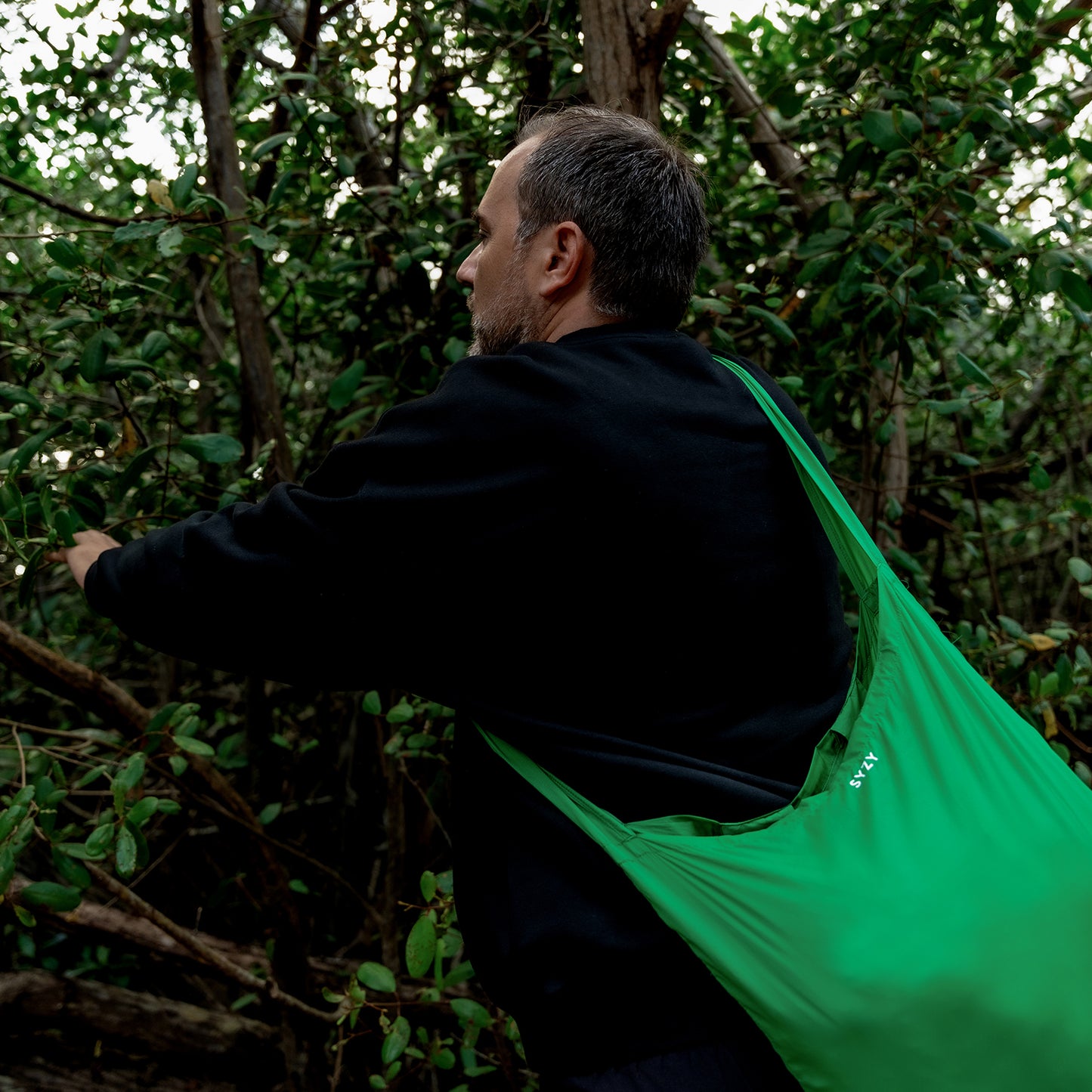 A man wearing a black sweatshirt is in a forest, holding branches and carrying the SYZY Tetra Viridian Green Packable Tote Bag.
