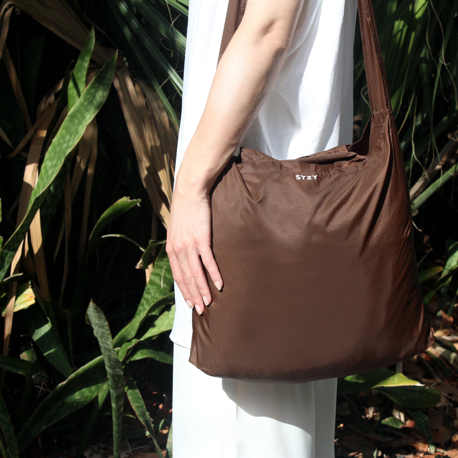 A person holding a Burnt Umber SYZY Tetra Tote Bag with multiple straps, surrounded by plants, against a natural background.