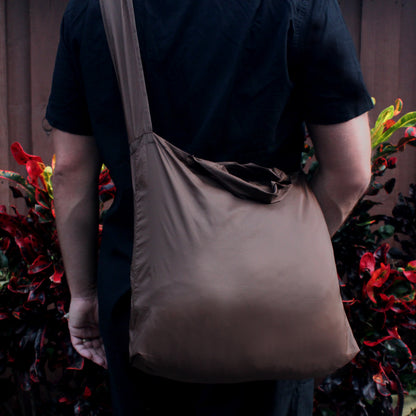 A person wearing a black shirt, carrying a Burnt Umber SYZY Tetra Tote Bag with a single shoulder strap, surrounded by colorful plants.