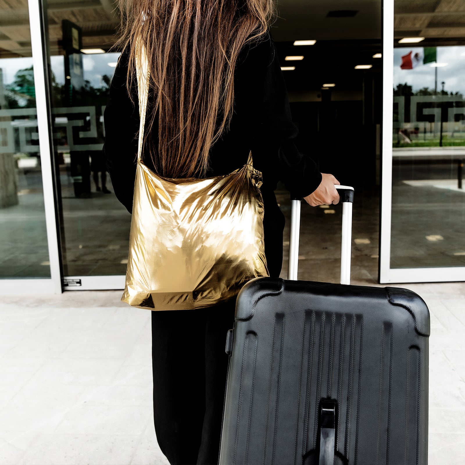 A woman holding a suitcase with a gold SYZY Tetra Metallic tote bag over her shoulder, walking toward a building entrance.