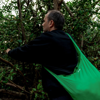A man wearing a black sweatshirt is in a forest, holding branches and carrying the SYZY Tetra Viridian Green Packable Tote Bag.