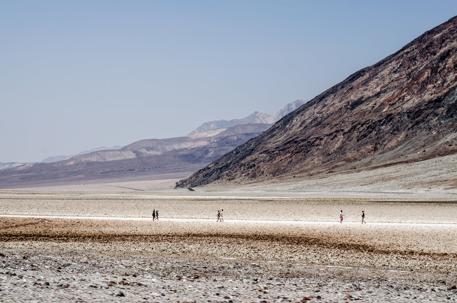 Vast desert landscape with mountains in the background, and two people walking along a dry, cracked path.