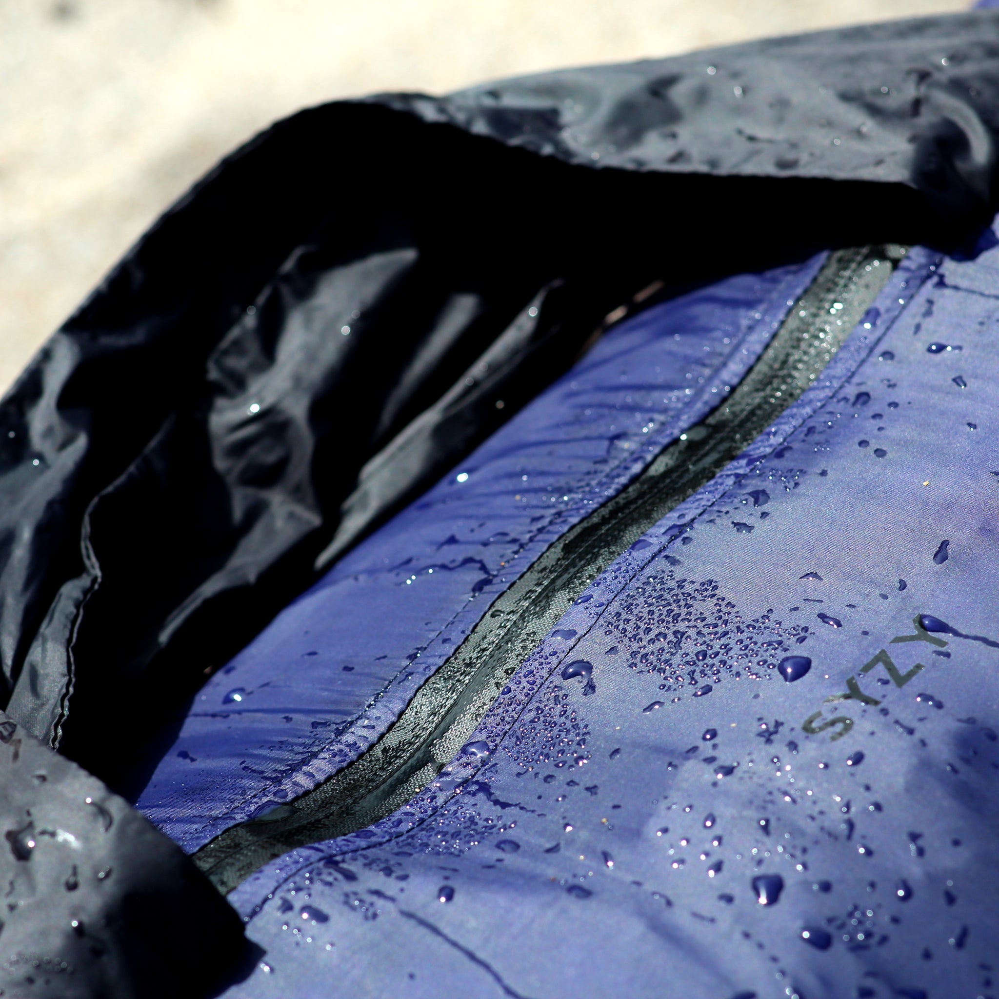 Close-up of a Rhom indigo SYZY bag with water droplets on the fabric, showing the zipper and logo.